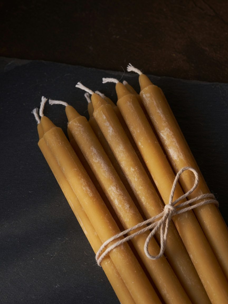 A bundle of unlit honey-gold beeswax tapers tied with twine on dark slate, with a pale film of bloom on the wax.